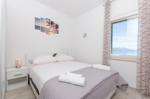 A neatly made bed with a grey quilt, white pillows, and rolled towels on a white nightstand. A wall-mounted artwork depicts a beach scene. - Apartment Paradise (Postira)