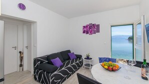 Ein modernes Wohnzimmer mit einem schwarz-weißen Couchgarnitur, einem Esstisch mit Obst und Weingläsern sowie Meerblick durch ein großes Fenster.