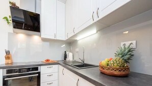 Une cuisine moderne avec des meubles blancs, un four encastré, un plan de travail noir et un panier de fruits contenant des ananas.
