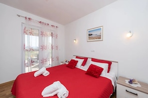 A hotel room with a large bed, red bedding, a window with sheer curtains, a bedside table with a lamp, and a framed picture on the wall. - House Ruza (Privlaka)