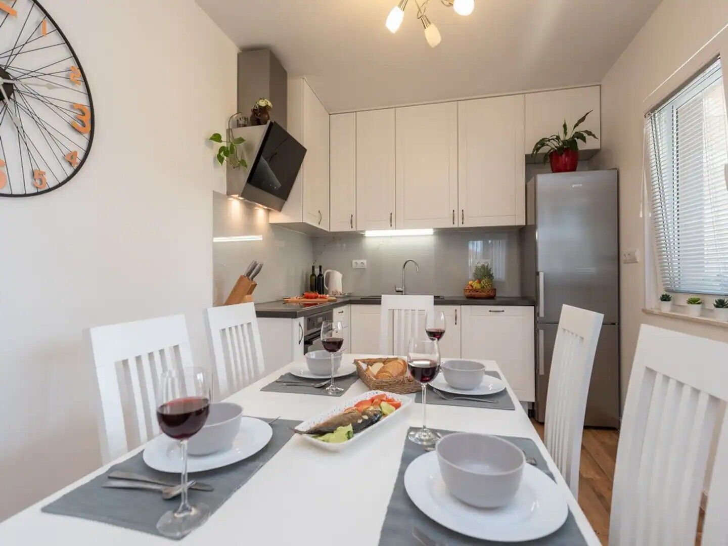 A modern kitchen with white cabinets, a stainless steel refrigerator, and a dining table set for two with wine glasses and a plate of food.