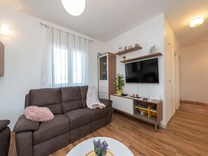A modern living room with a brown sofa, a flat-screen TV mounted on a wooden shelf, and a small table with a vase. - House Ruza (Privlaka)