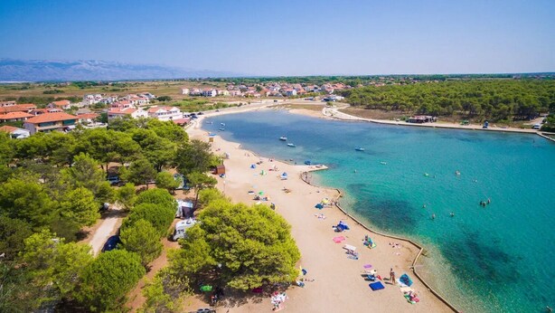 Vue aérienne d’une zone côtière avec une plage de sable, des eaux d’un bleu limpide et, en arrière-plan, des montagnes.