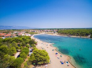 Vista aérea de uma área costeira com praia de areia, águas azuis cristalinas e montanhas ao fundo.