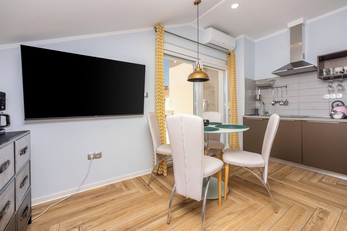 A modern kitchen with a dining area, featuring a flat-screen TV, a dining table with chairs, and a kitchen counter with utensils.