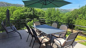 Een terras buiten met een tafel en stoelen, een groene parasol en uitzicht op groene bomen en bergen.