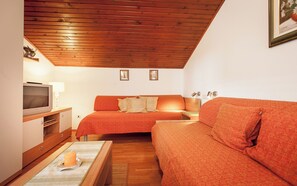 Ein gemütliches Zimmer mit Holzbalkendecke, zwei Betten mit orangefarbener Bettwäsche, einem Fernseher und einem kleinen Tisch mit einer Kerze.