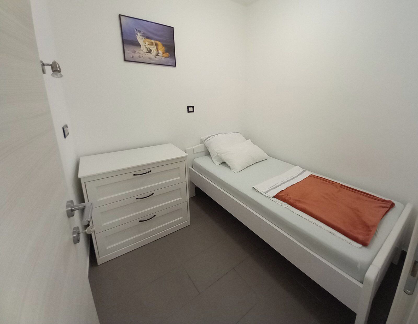 A small, white, single-bed room with a bedside table and a drawer unit.