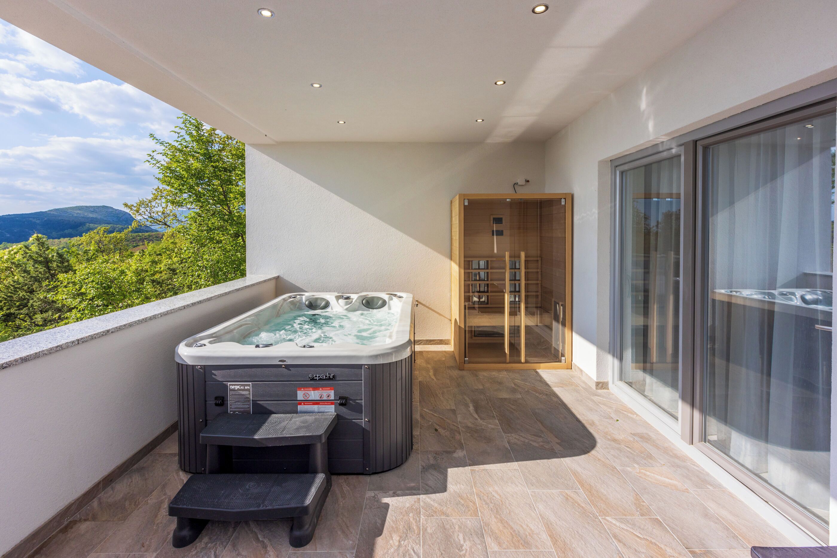 A balcony with a hot tub, a glass-enclosed shower area, and a view of greenery and hills.
