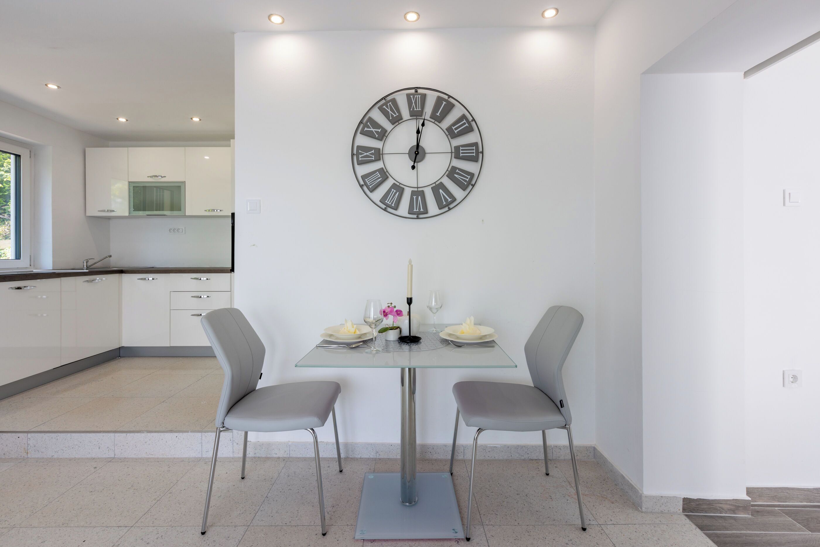 A modern kitchen with white cabinets, a glass dining table, two grey chairs, and a large wall clock.