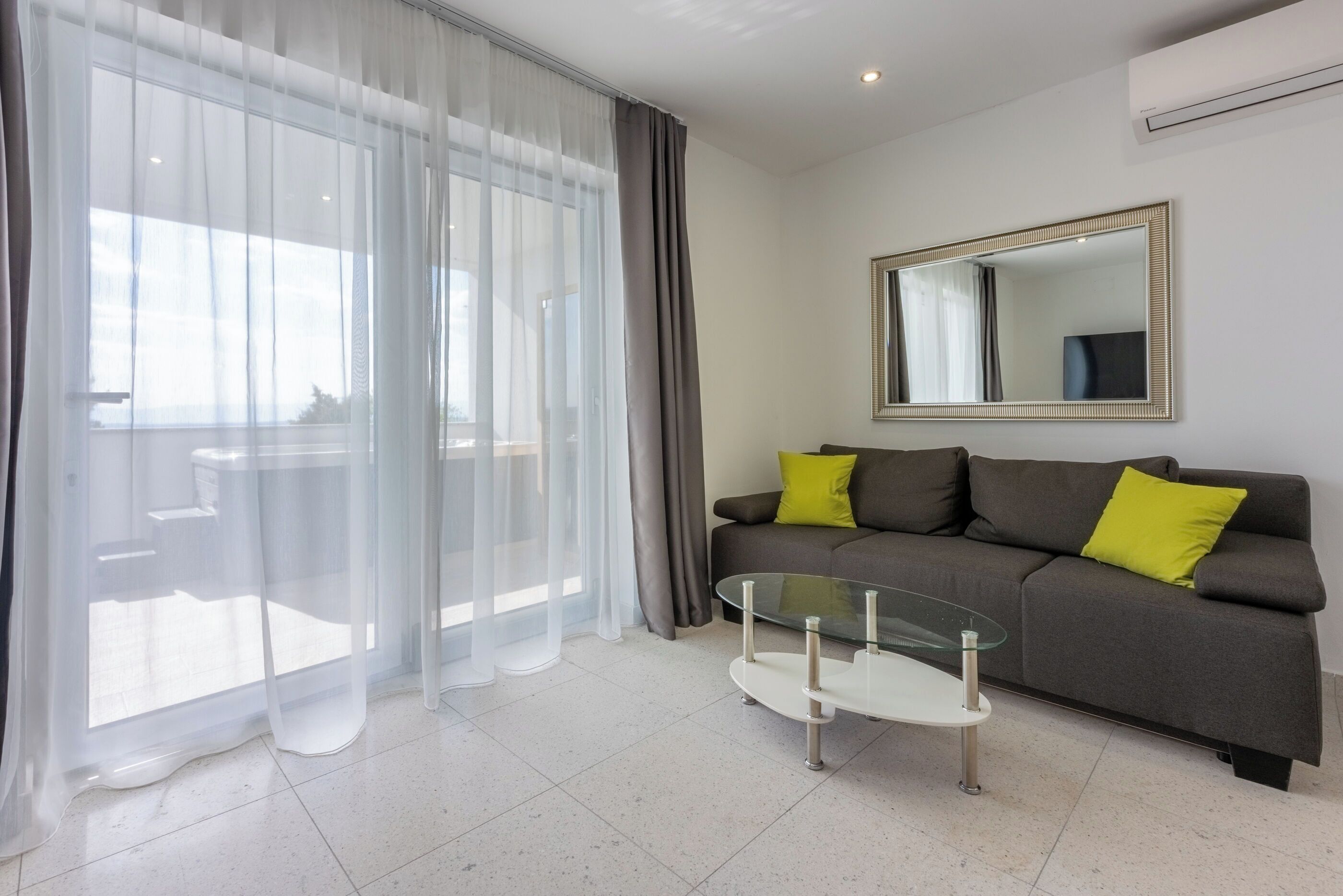 A modern living room with a grey sofa, two yellow cushions, a glass coffee table, sheer curtains, and a large mirror on the wall.