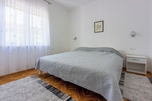 Un dormitorio con cama, mesita de noche, lámpara de pared y ventana con cortinas translúcidas.