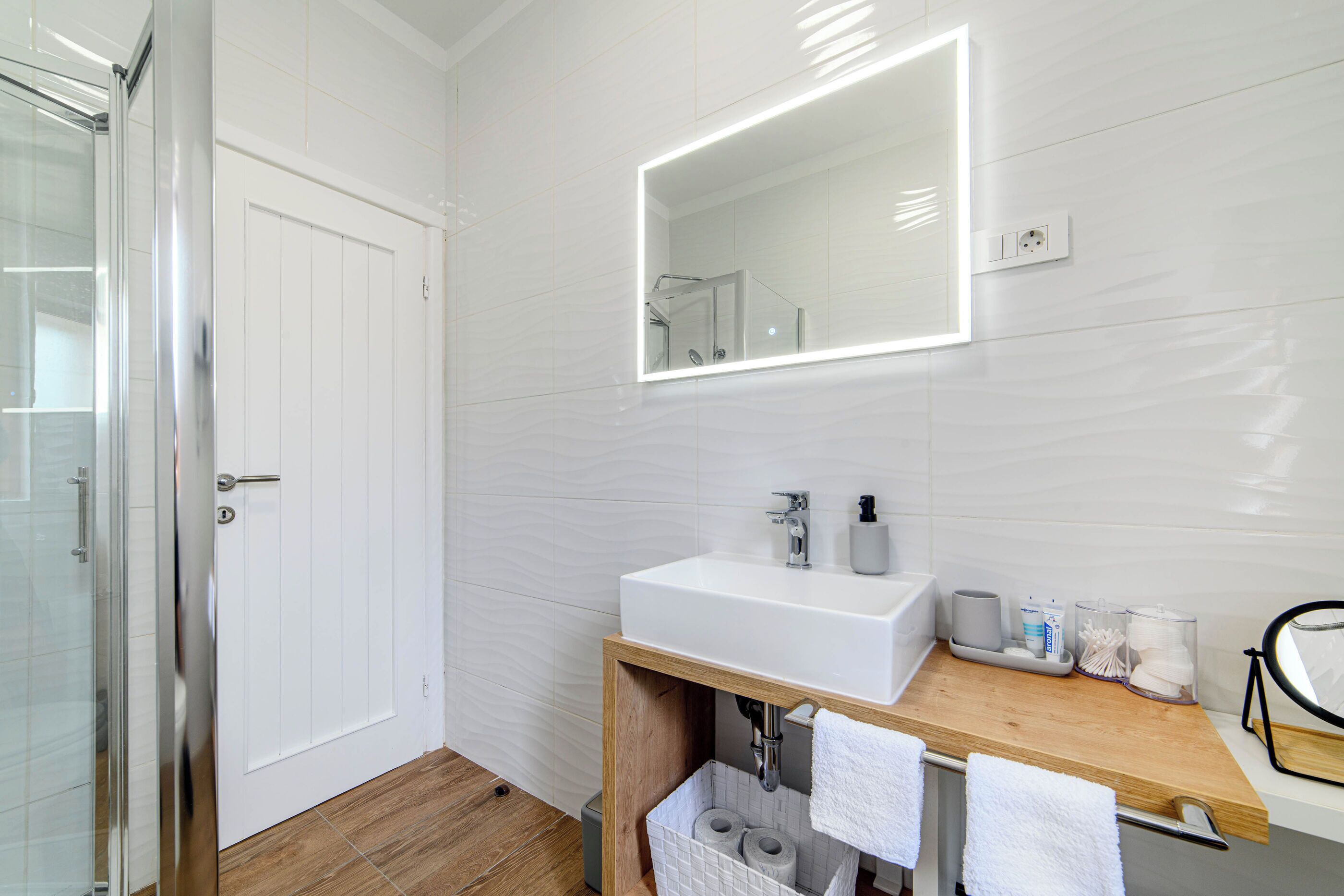 Une salle de bain moderne avec une douche à l’italienne en verre, un lavabo blanc avec un miroir au-dessus, et un meuble-lavabo en bois agrémenté de serviettes et d’articles de toilette.