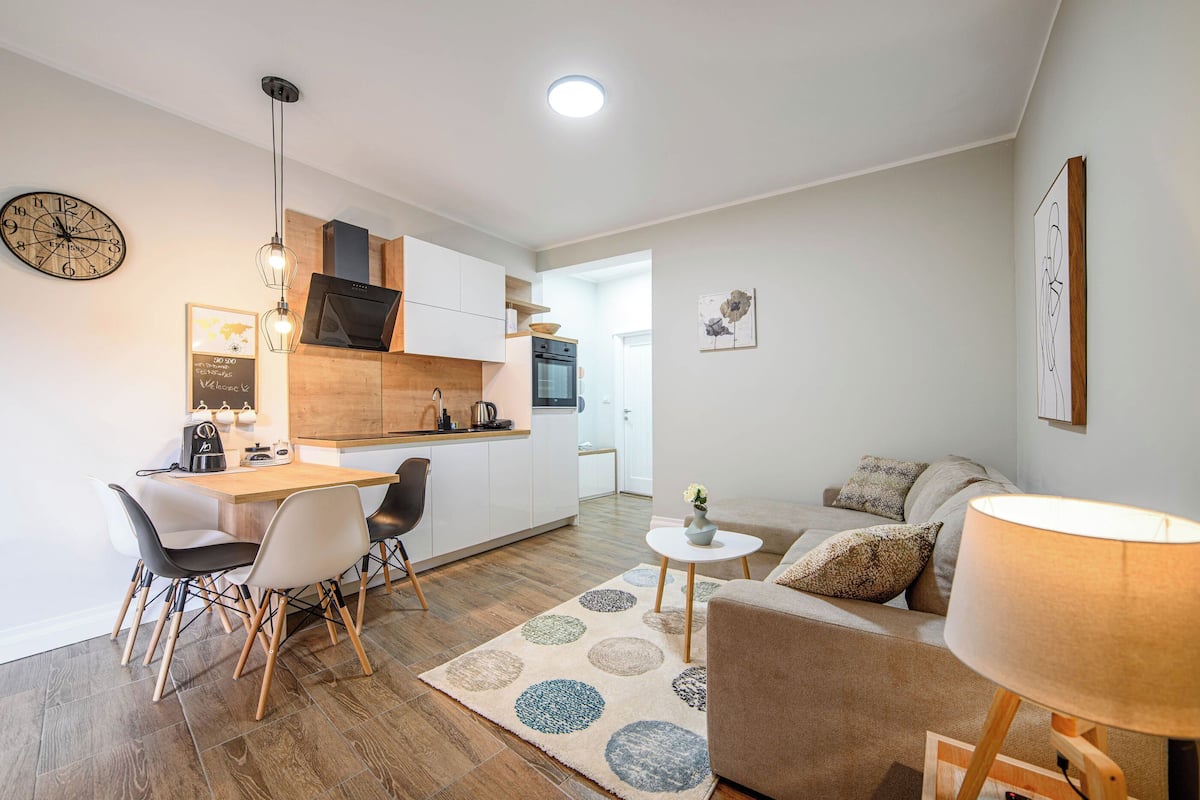 A modern apartment with a kitchen, dining area, and living room. It includes a wooden floor, white walls, and a large clock.