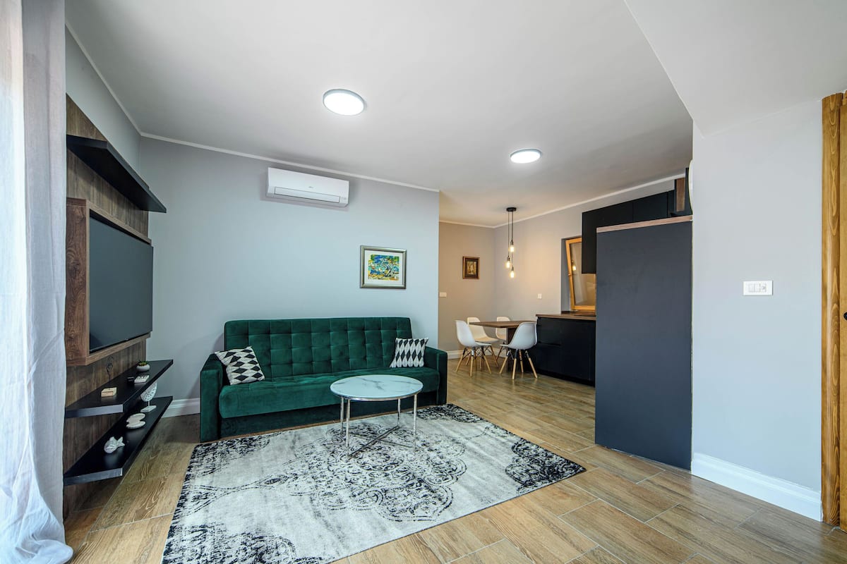 A living room with a green sofa, a small round coffee table, and a patterned rug. There is a flat-screen TV on a wooden stand, a dining area with a table and chairs, and a kitchen area with a refrigerator and cabinets.