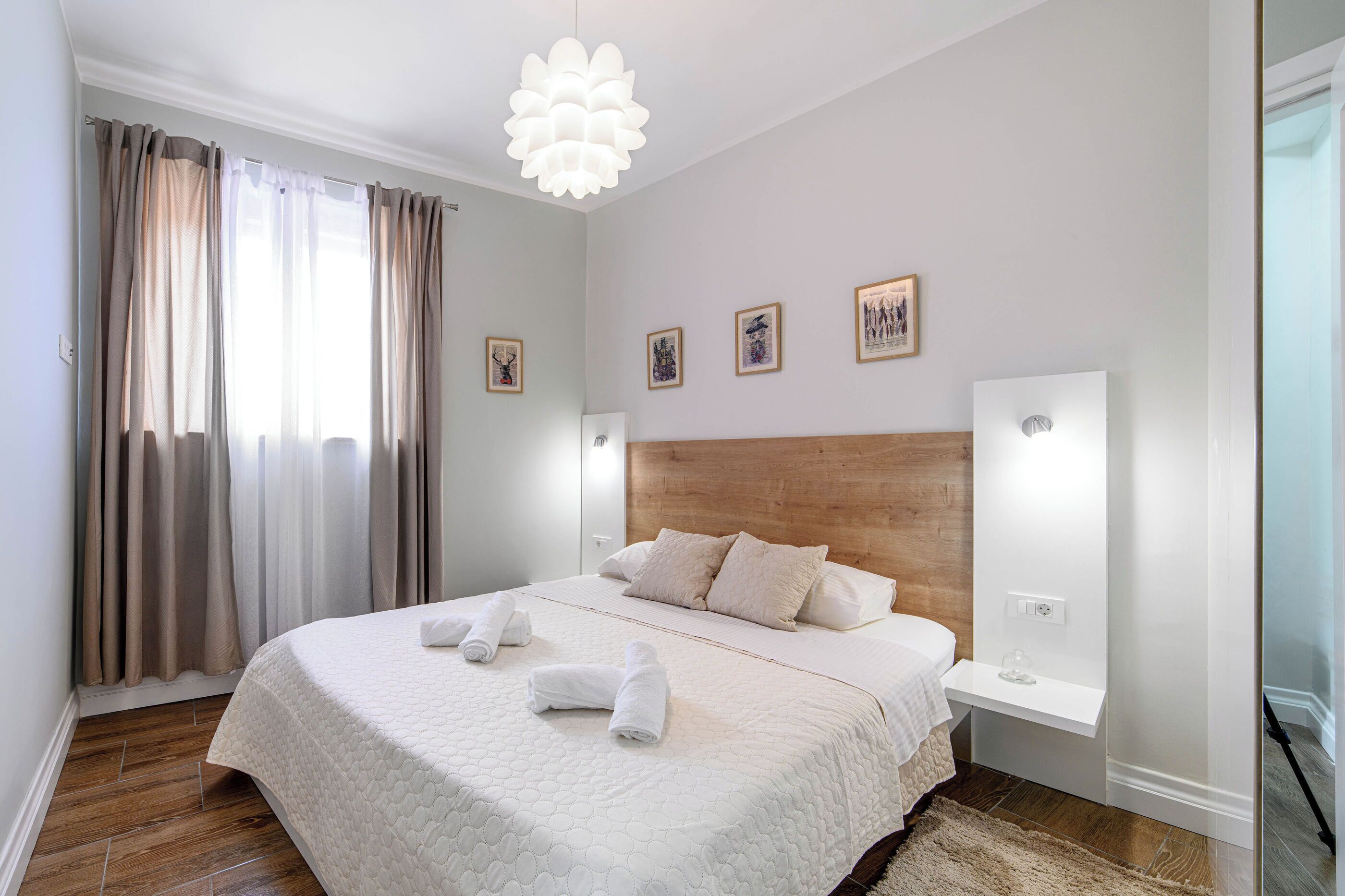 A neatly made bed with a wooden headboard, a bedside table with a lamp, and a large window with curtains.