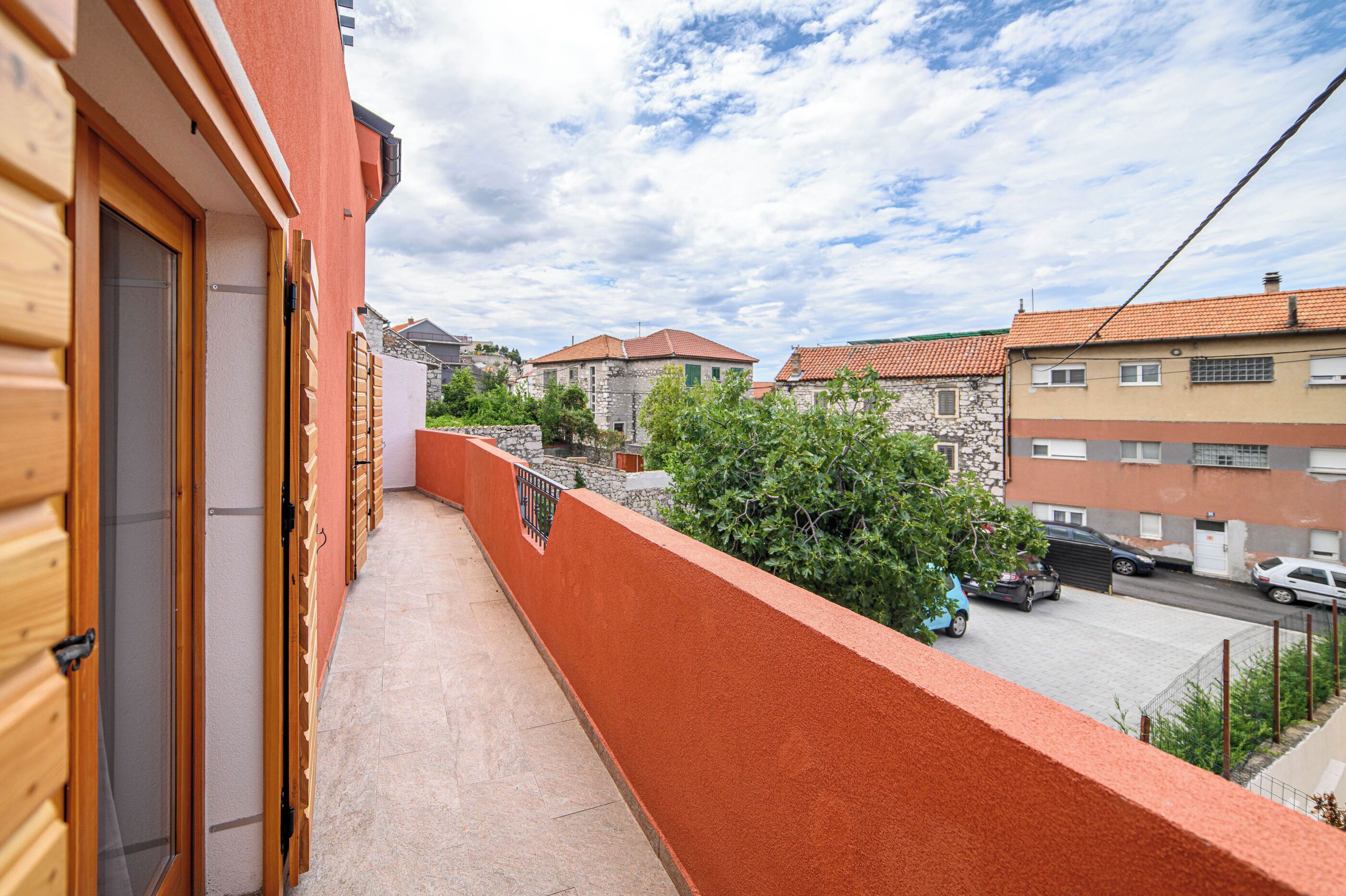 Un balcón con un muro rojo, una puerta de madera y vistas a edificios vecinos y coches aparcados.