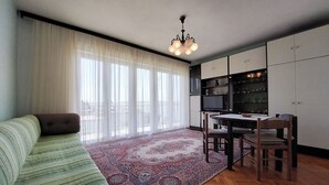 En moderne stue med sofa, spisebord med stole, et køkkenområde med skabe og et stort vindue med lette gardiner.