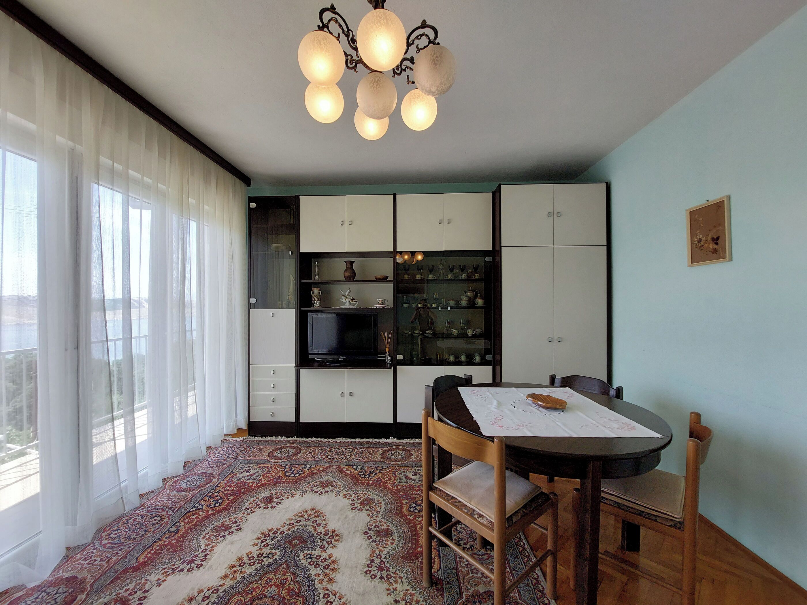 A dining area with a table, chairs, a patterned rug, and a cabinet with shelves containing dishes and a television.