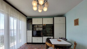 A dining area with a table, chairs, a patterned rug, and a cabinet with shelves containing dishes and a television. - Apartment Vlasta (Crikvenica)