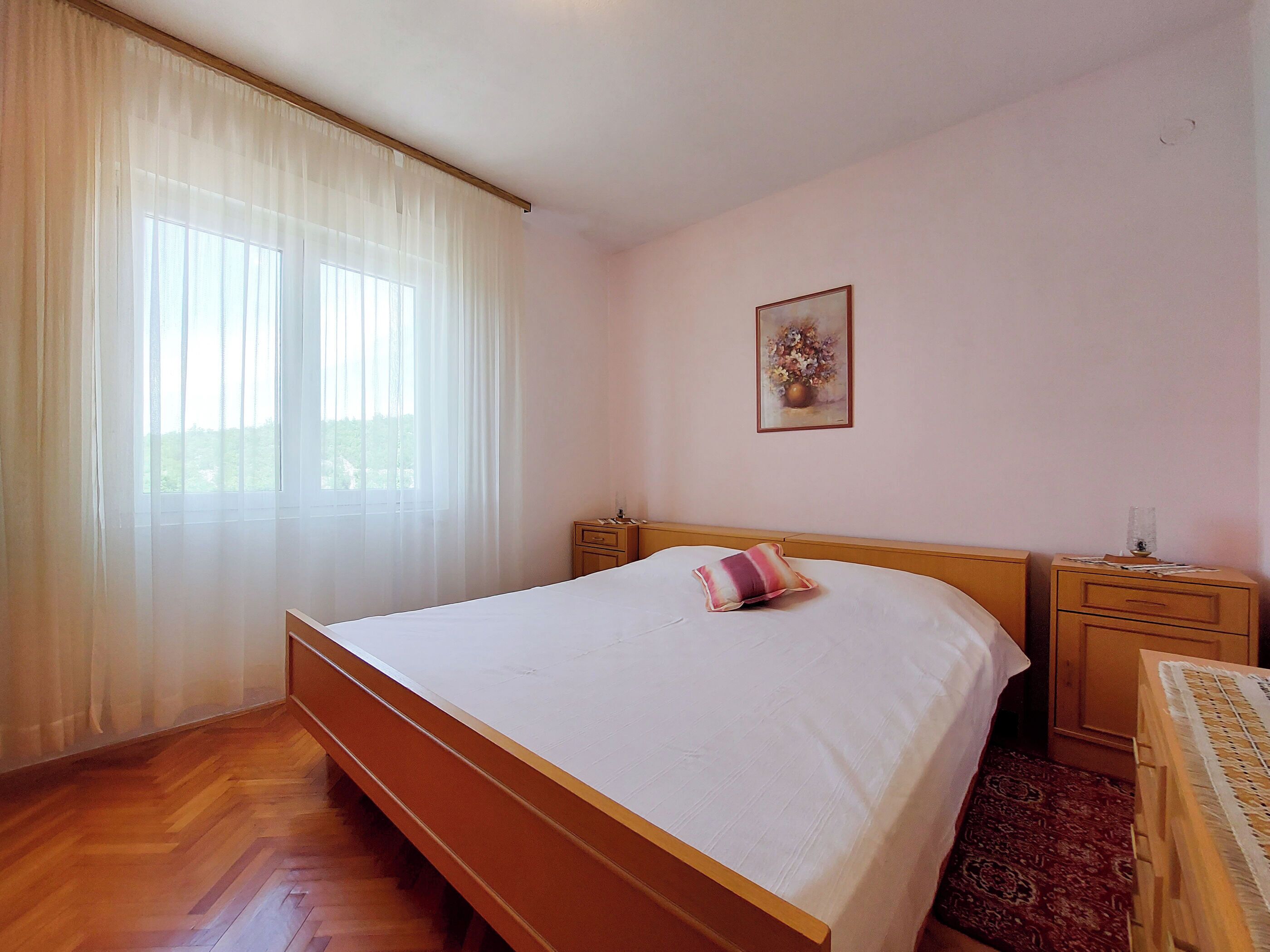 Un dormitorio con cama de madera, una mesita de noche y una ventana con cortinas ligeras.
