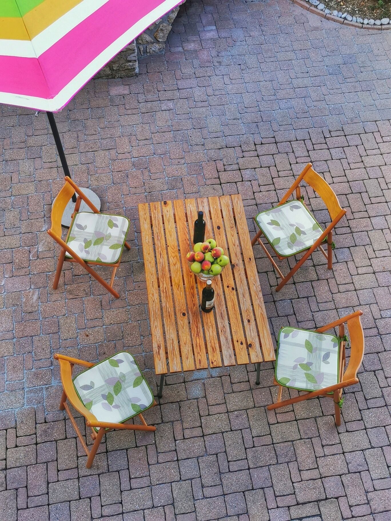 Una mesa de madera con cuatro sillas, cada una con un cojín estampado con motivos de hojas, rodeada de un pavimento de ladrillo.