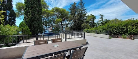 An outdoor patio with a table and chairs, surrounded by greenery and a clear sky.