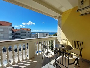 Un balcón con mesa y sillas, con vistas a una zona costera con edificios y un cielo azul despejado.