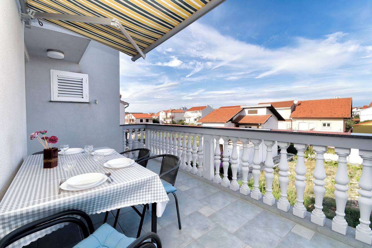 Een balkon met een geruit tafeltje gedekt voor een maaltijd, witte hekwerkleuningen en een uitzicht op gebouwen met rode daken op de achtergrond.
