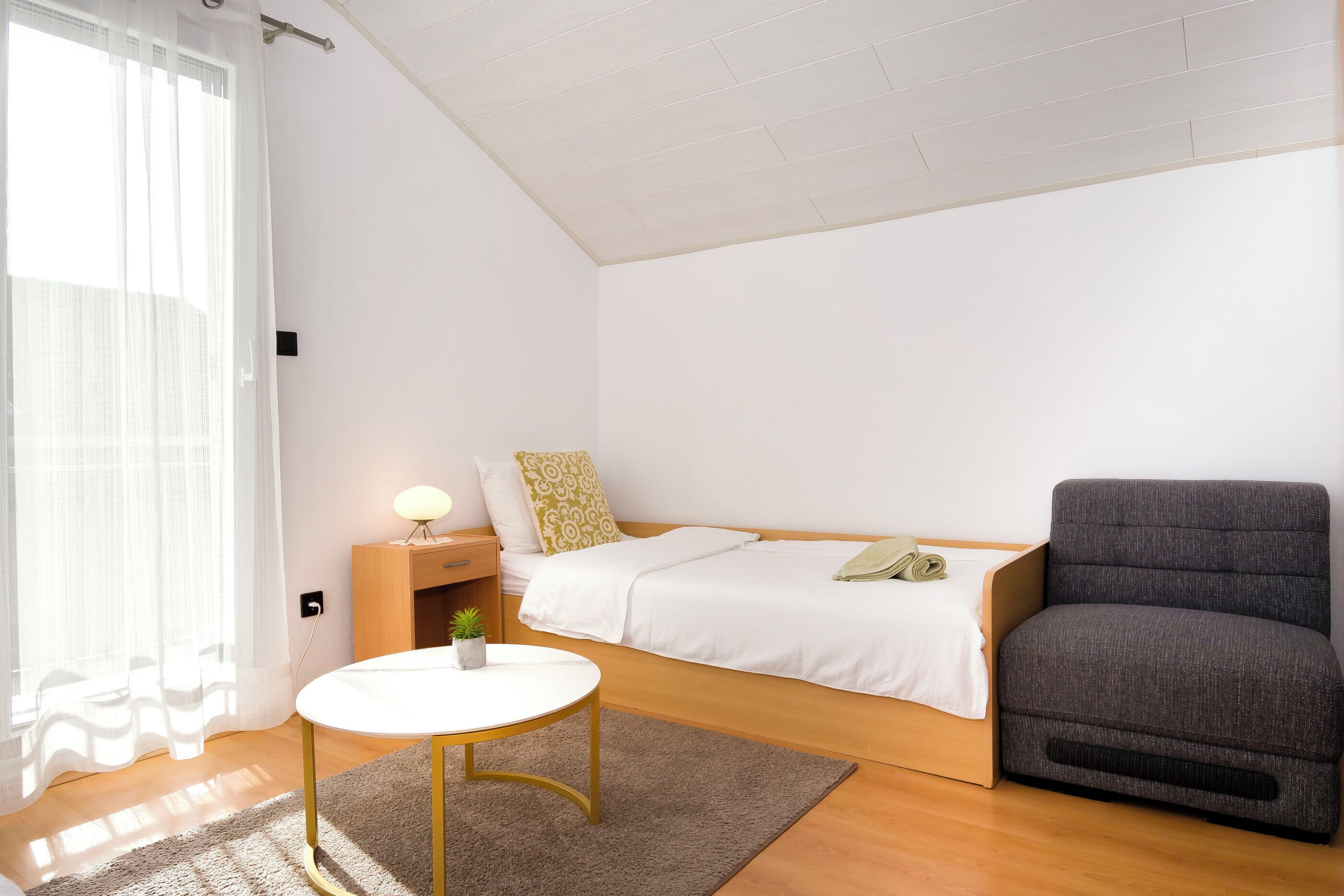 A compact bedroom with a bed, bedside table, lamp, and a grey armchair.