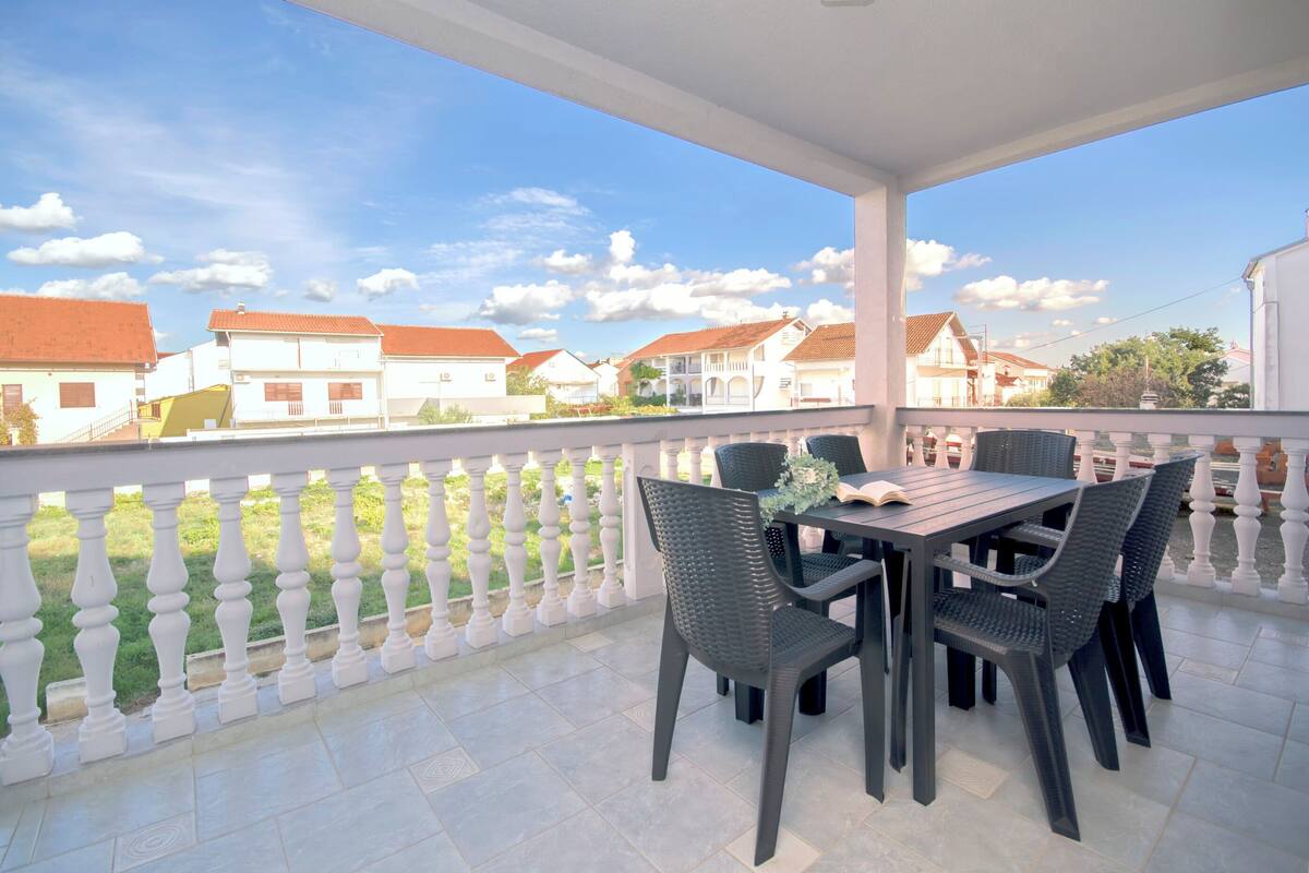 Een overdekt balkon met een tafel en stoelen, een witte balustrade en een uitzicht op huizen in de verte.