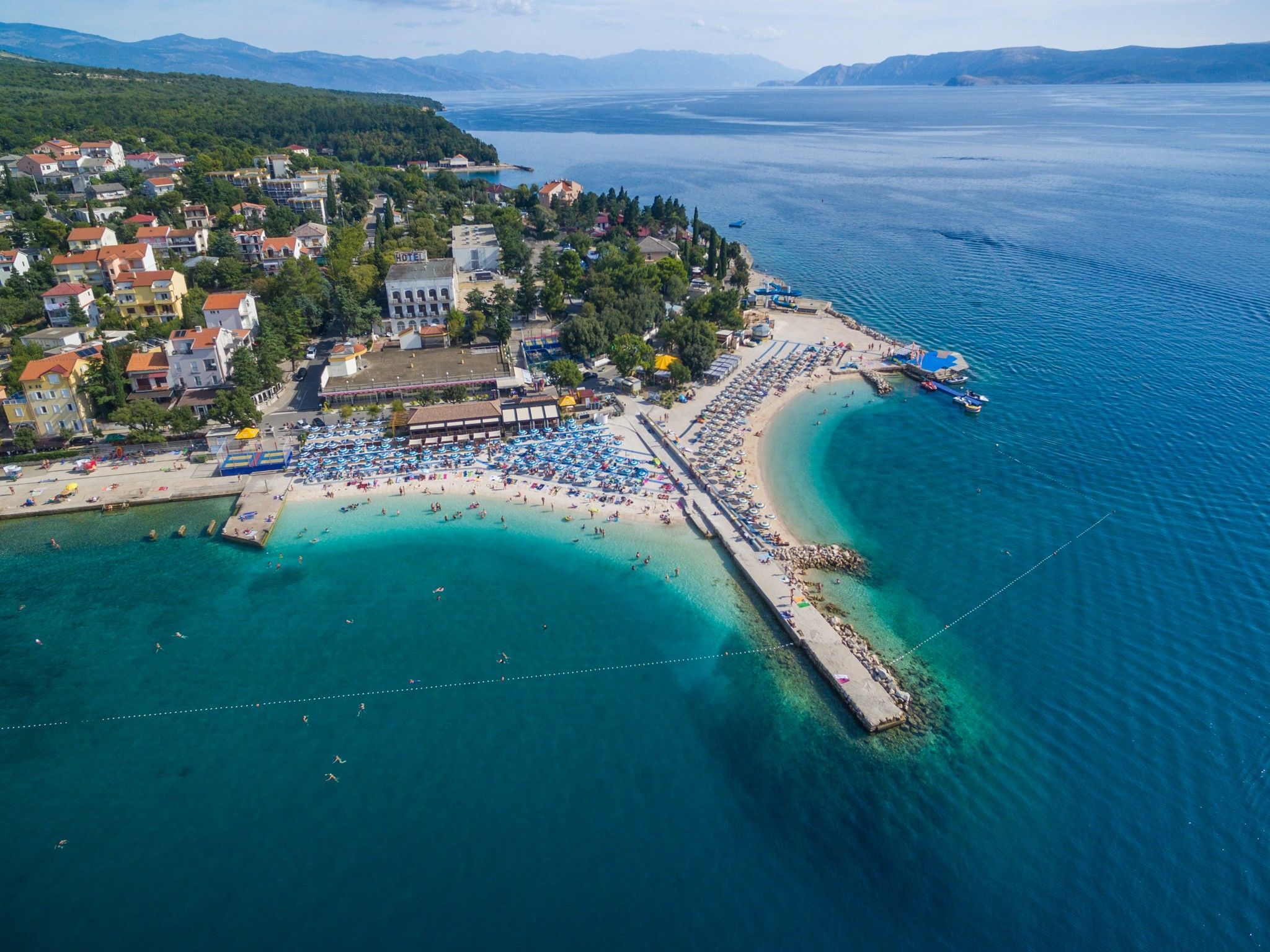 An aerial view of a coastal town with a beach, a hotel, and a pier extending into the sea.