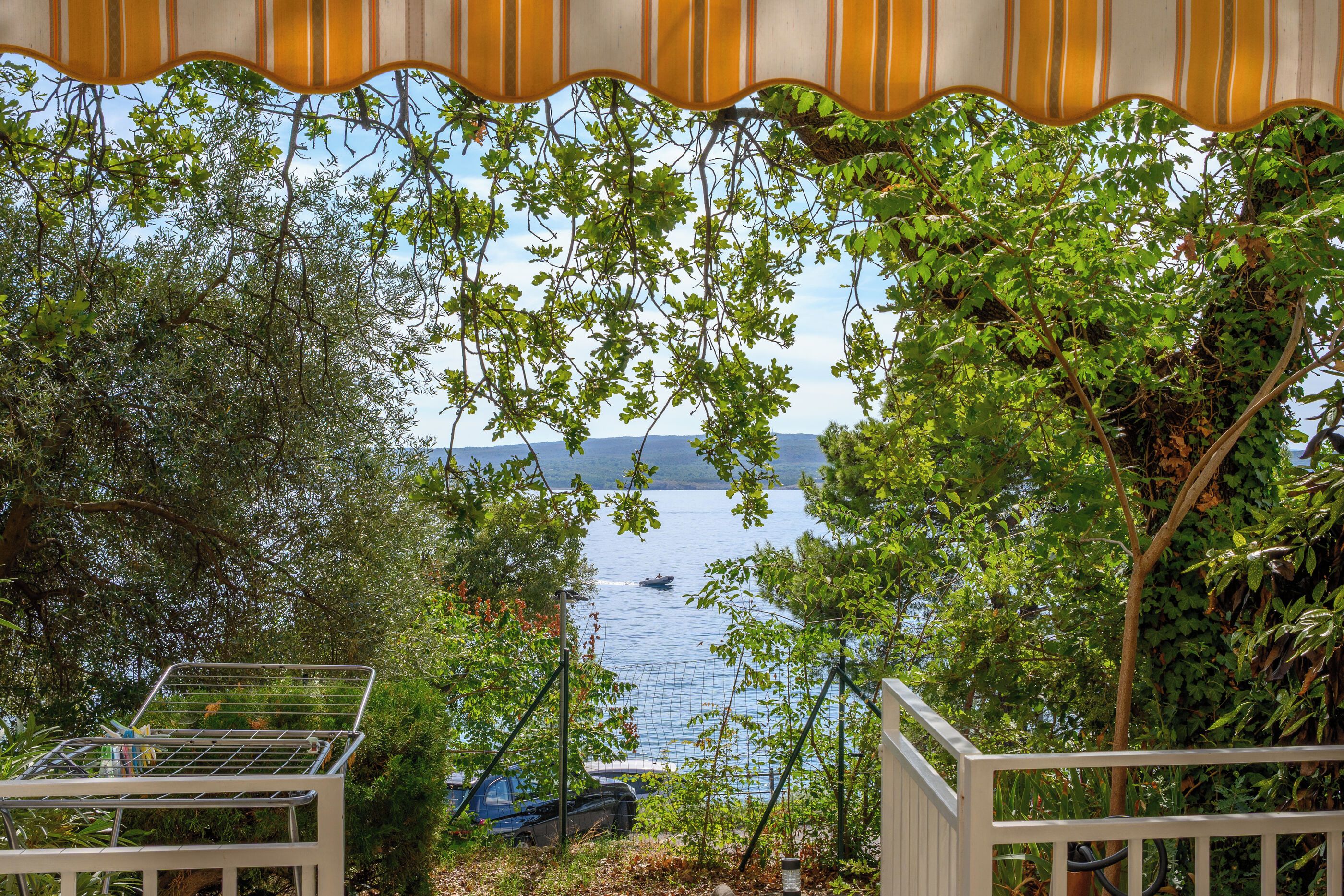 A view through a garden with a metal fence, a boat on the water, and a car parked nearby.