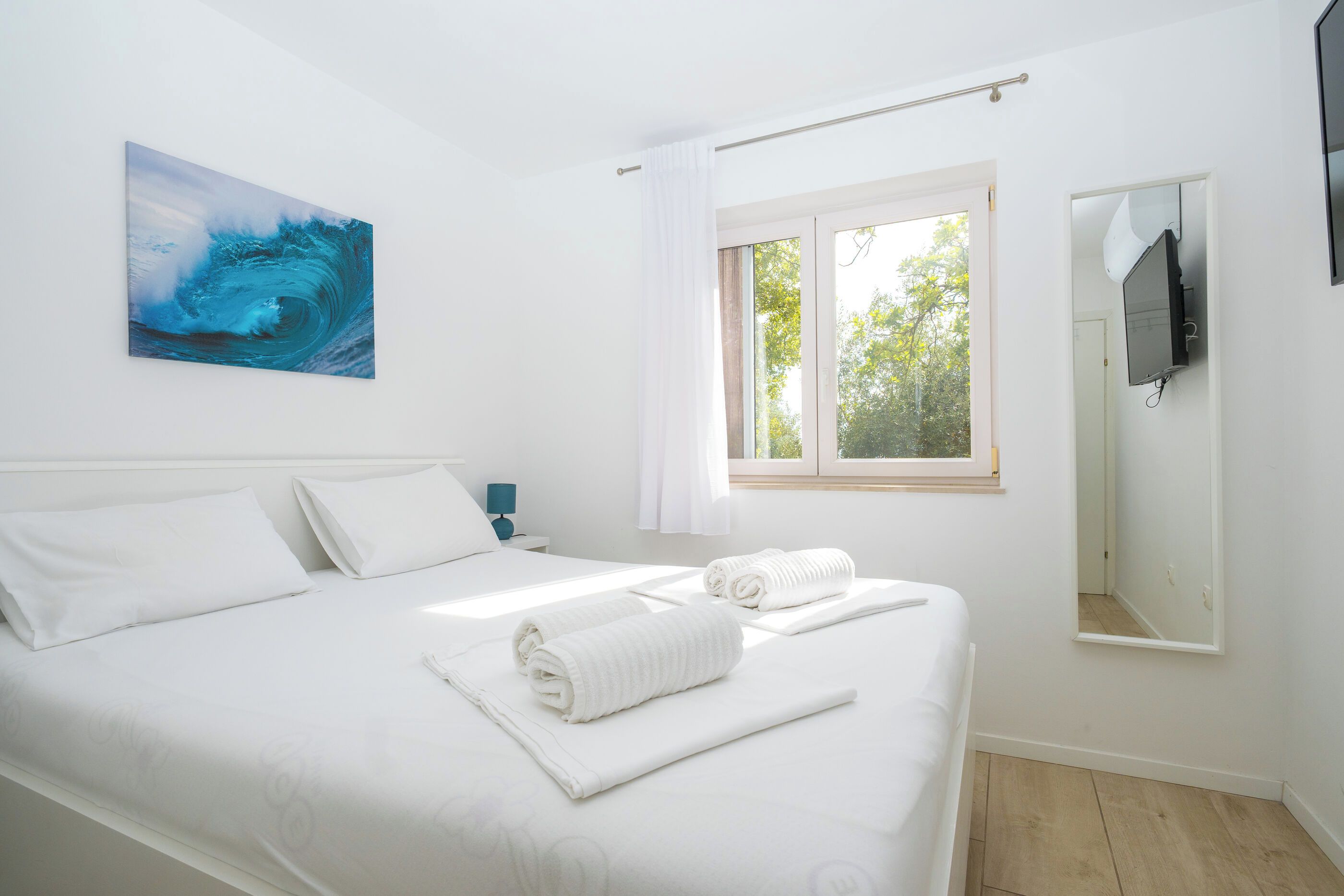 A hotel room with a bed, a window with curtains, a flat-screen TV mounted on the wall, and a painting of a wave on the wall.