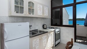 Cucina compatta con frigorifero, fornello e lavello. Sono presenti armadi con ante in vetro e una finestra con vista sul mare.