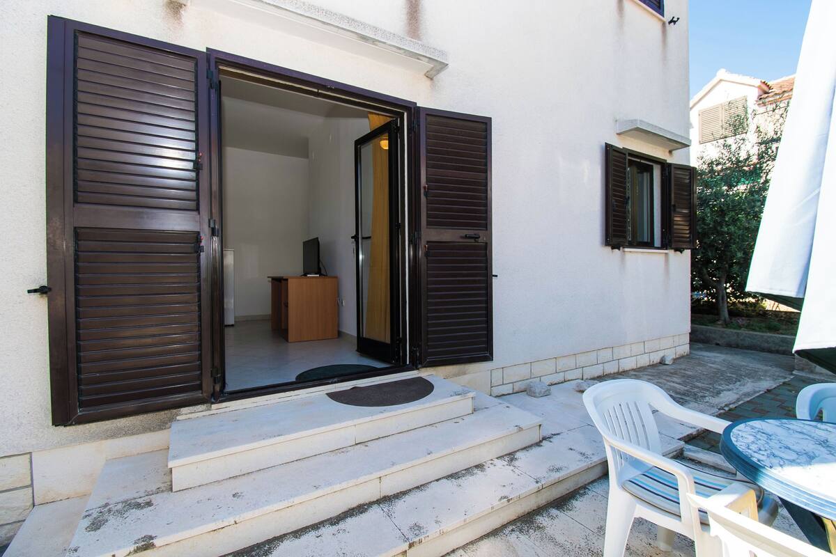 A residential property with a sliding glass door entrance, a small wooden cabinet, and a white plastic chair and table outside.