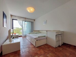 Quarto com cama, uma pequena escrivaninha, radiador, janela com cortinas e varanda.