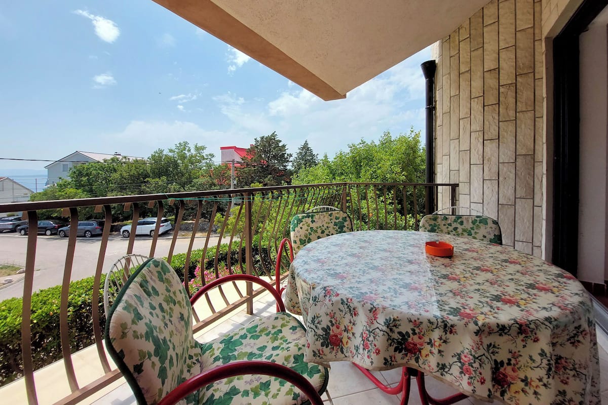 A balcony with a round table and chairs, a view of a parking lot, and greenery in the background.