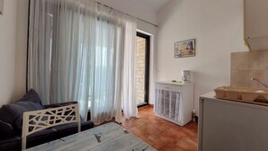 A small, well-lit room with a dining table, a sofa, and a kitchen area with a coffee maker and storage containers. - Apartments Ilija (Crikvenica)