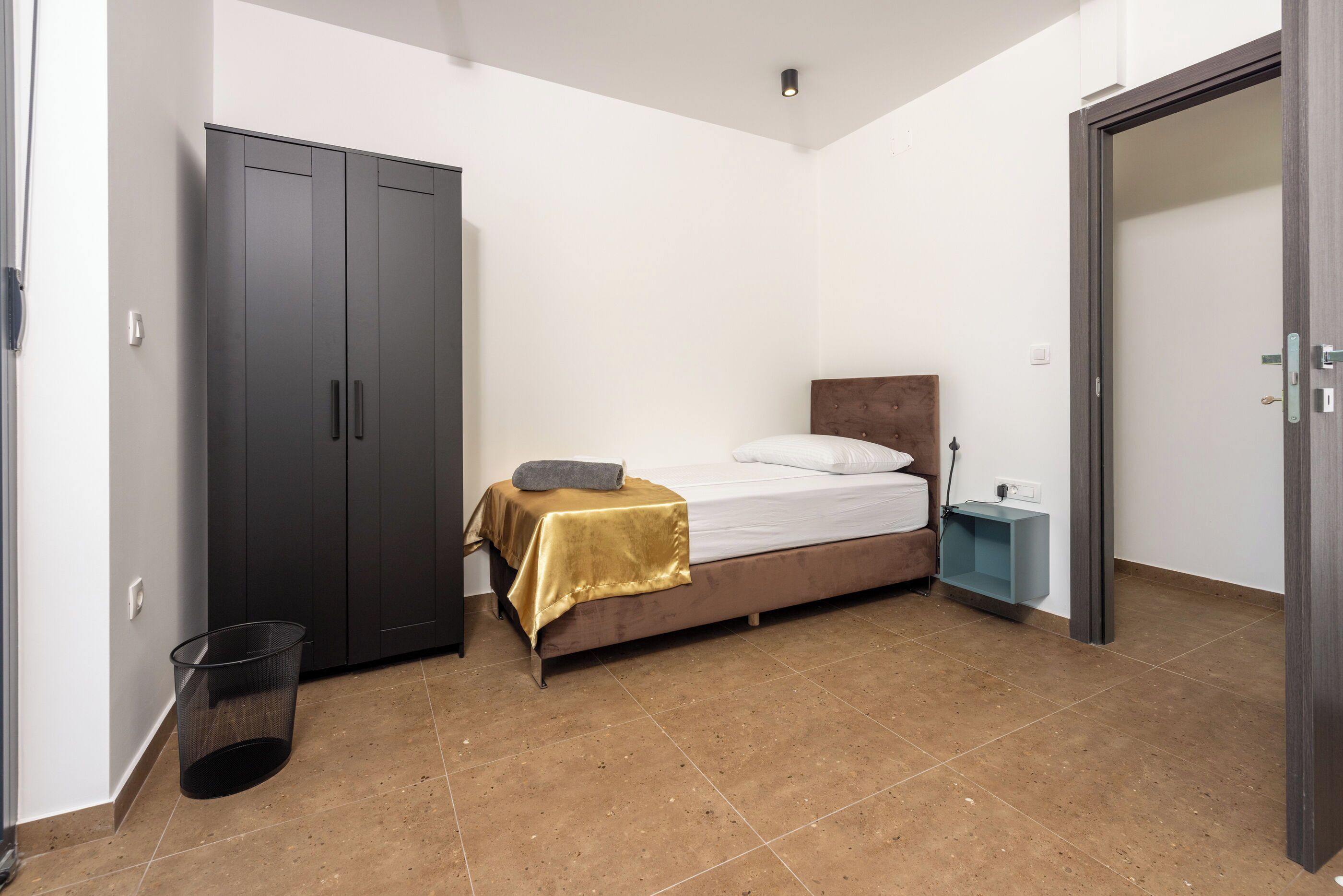 A bedroom with a bed, a wardrobe, a bedside table, and a trash bin.