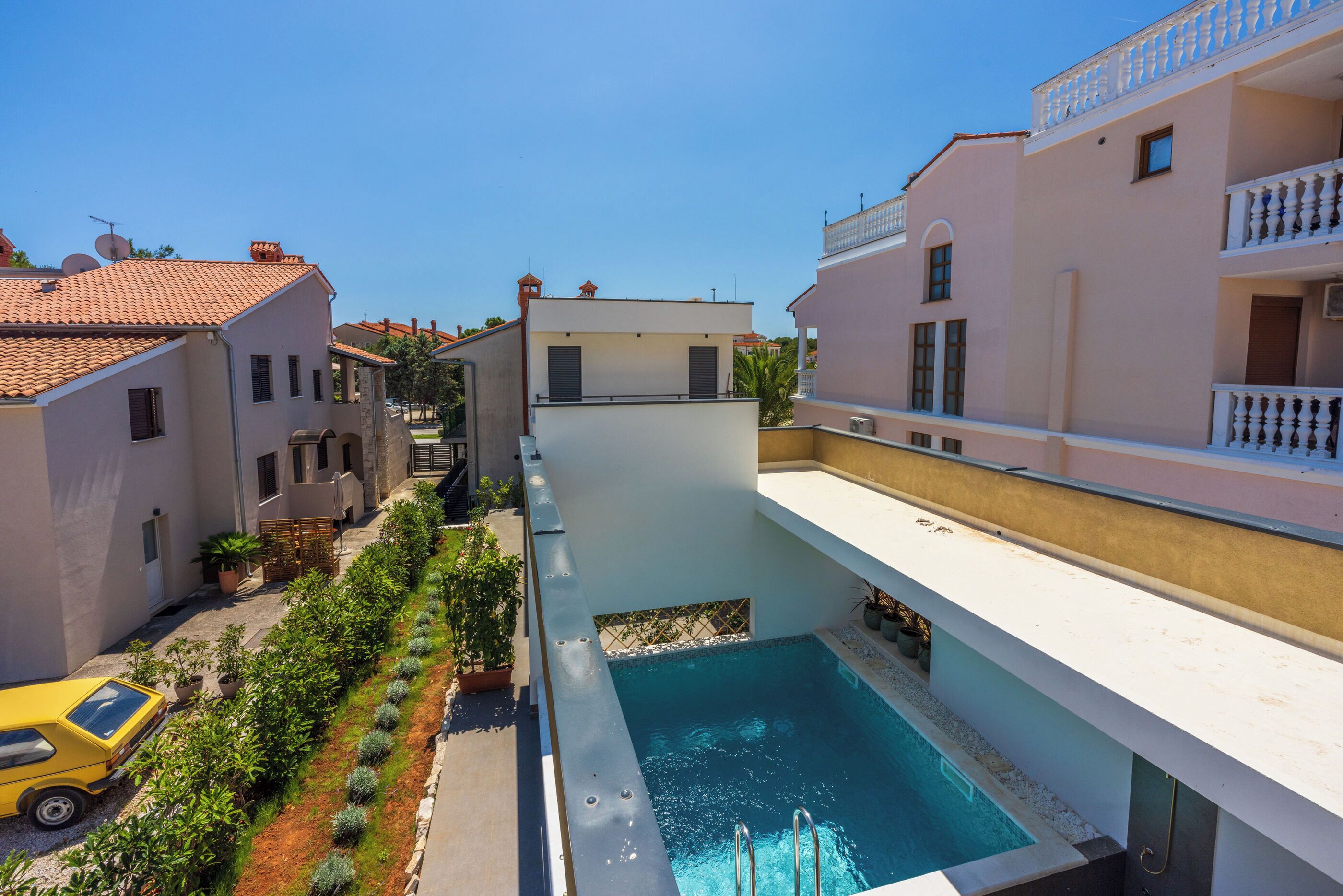A modern building with a swimming pool, a yellow car, and a row of plants along the side.