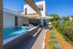 A modern outdoor pool area with a clear blue pool, a covered seating area, and a well-maintained garden. - House Neptunia (Medulin)