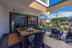 An outdoor patio with a wooden table and chairs, a glass door leading to an interior space, and a large umbrella. - House Neptunia (Medulin)