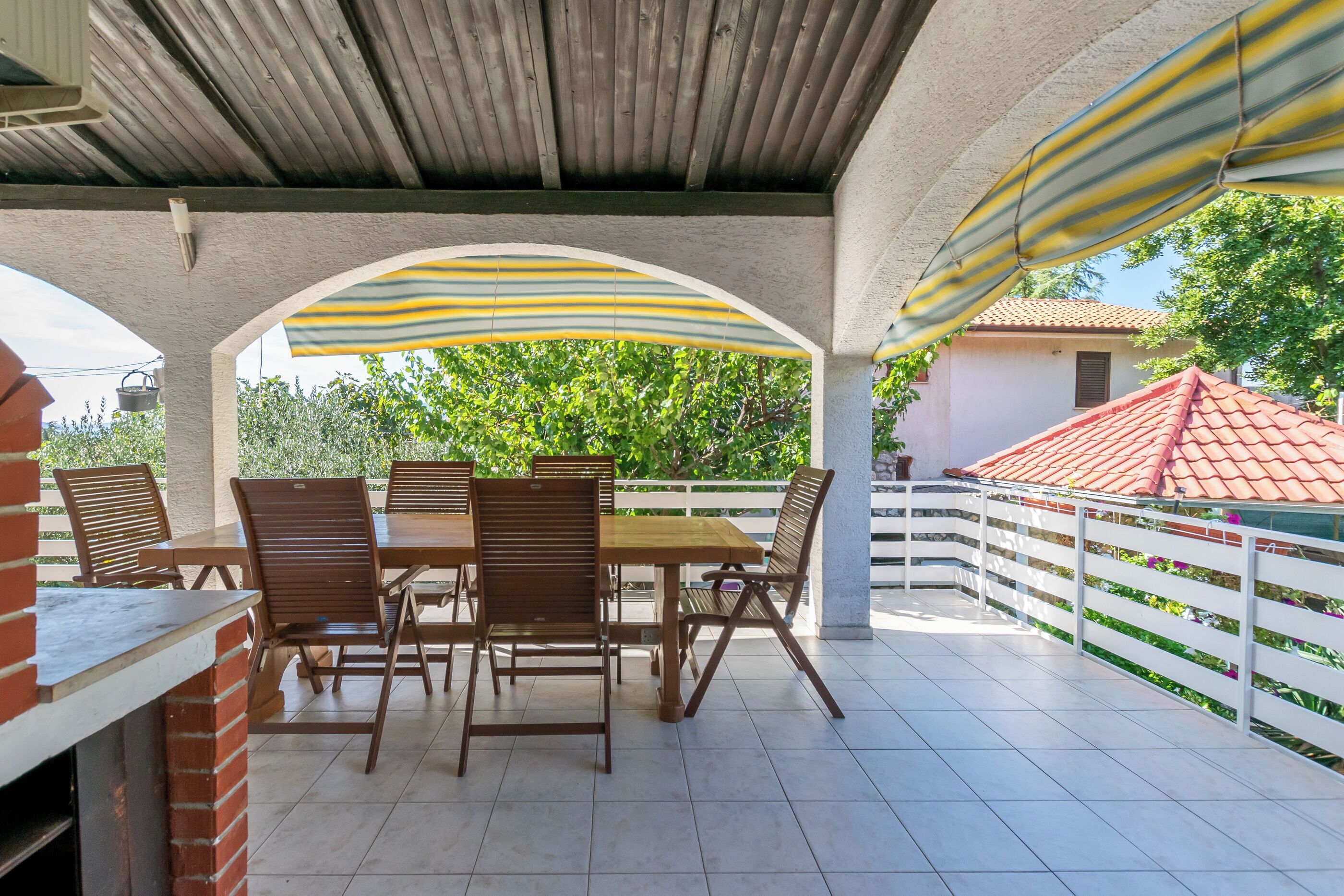 An outdoor patio with a brick fireplace, wooden table and chairs, and a striped awning providing shade.