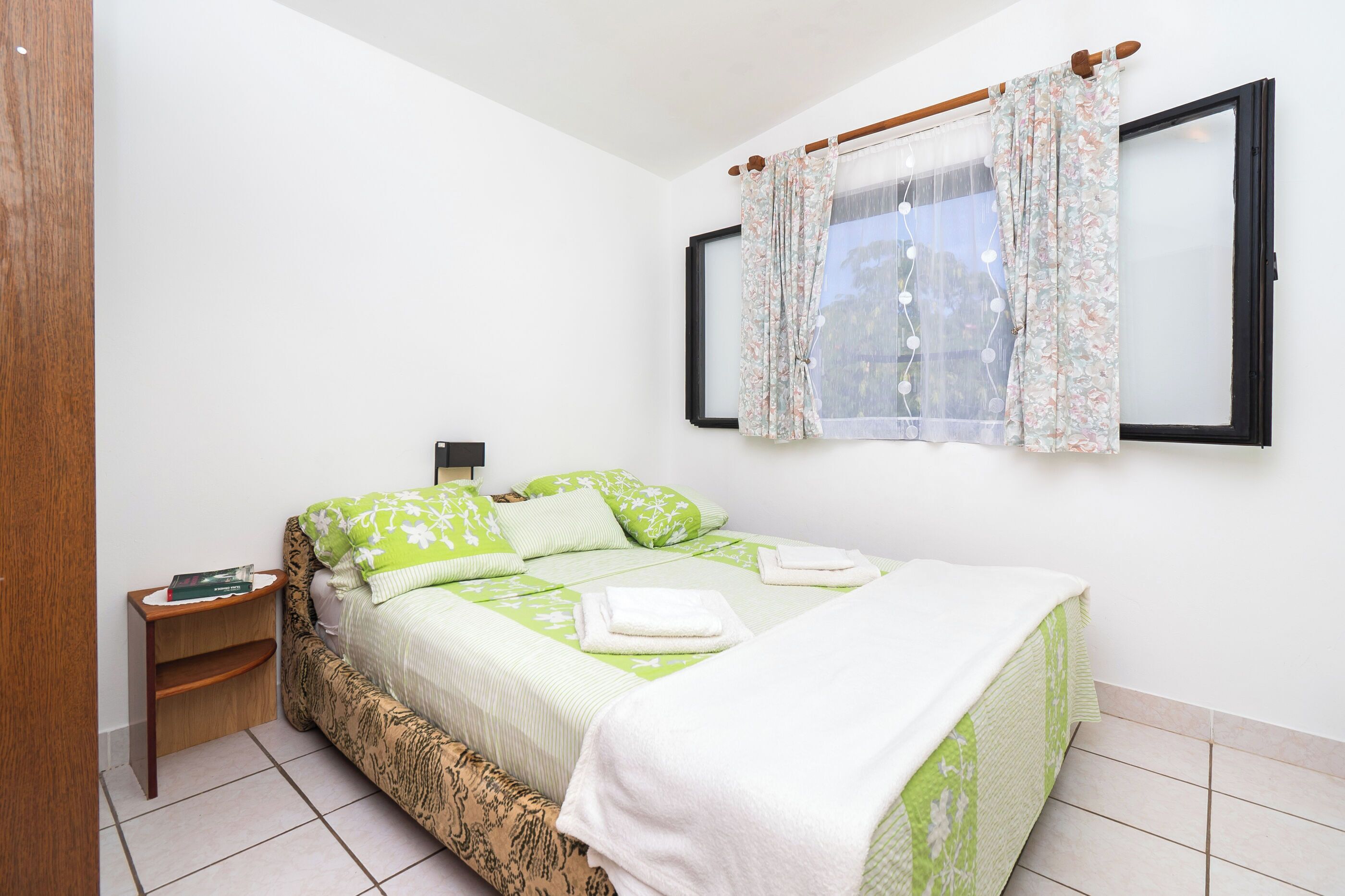A neatly made bed with green bedding, a bedside table with a book, and a window with floral curtains.
