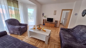 Ett vardagsrum med två soffor i mörkt mönster, ett vitt soffbord med frukt och en vas, en tv-apparat på ett träskåp och en balkong med utsikt.