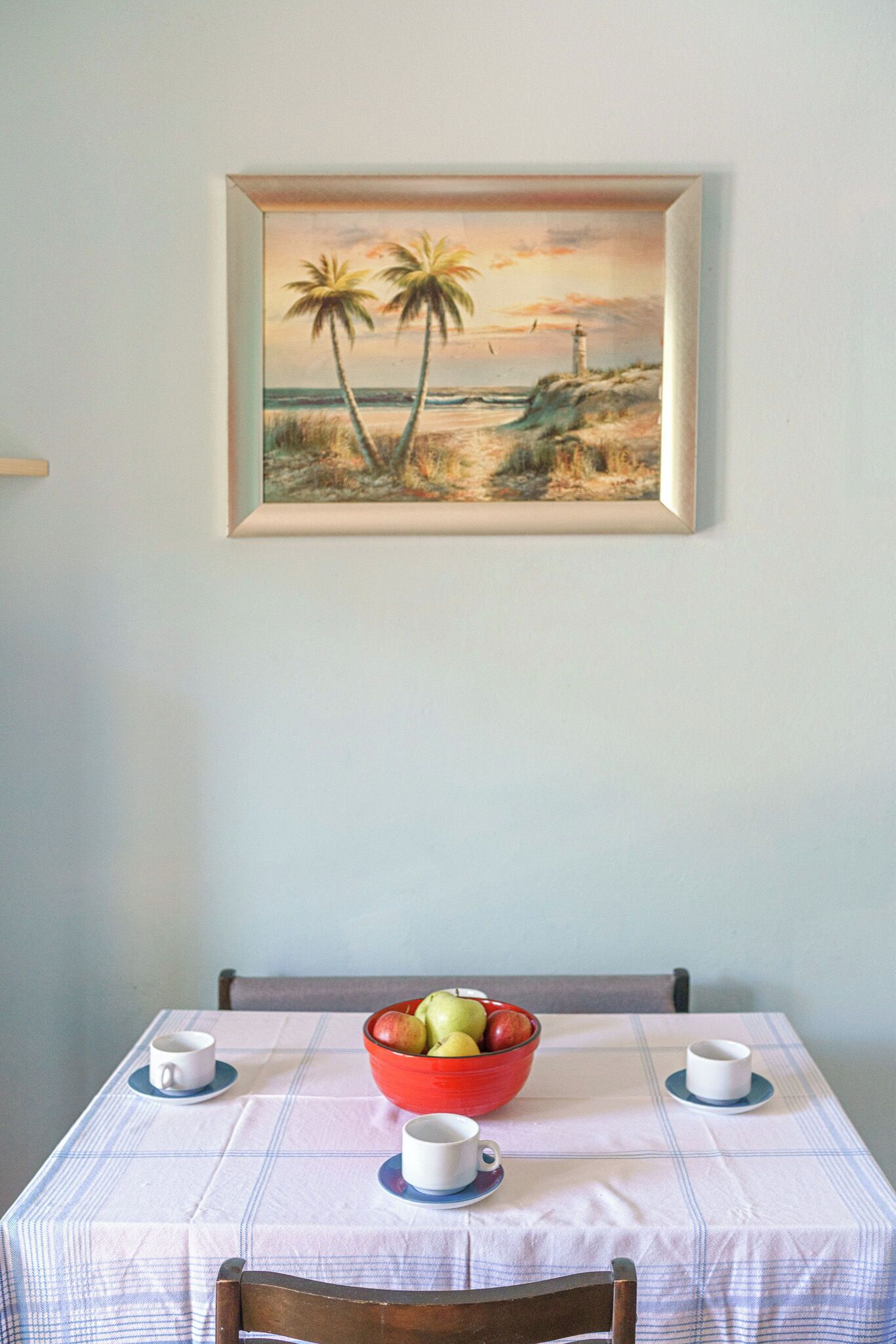 A dining table with a checkered tablecloth, a bowl of fruit, and two cups on a wall with a framed painting of palm trees and a lighthouse.