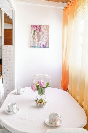 Une table ronde recouverte d’une nappe blanche, deux tasses blanches, un vase en verre contenant des fleurs roses et un bol de biscuits.