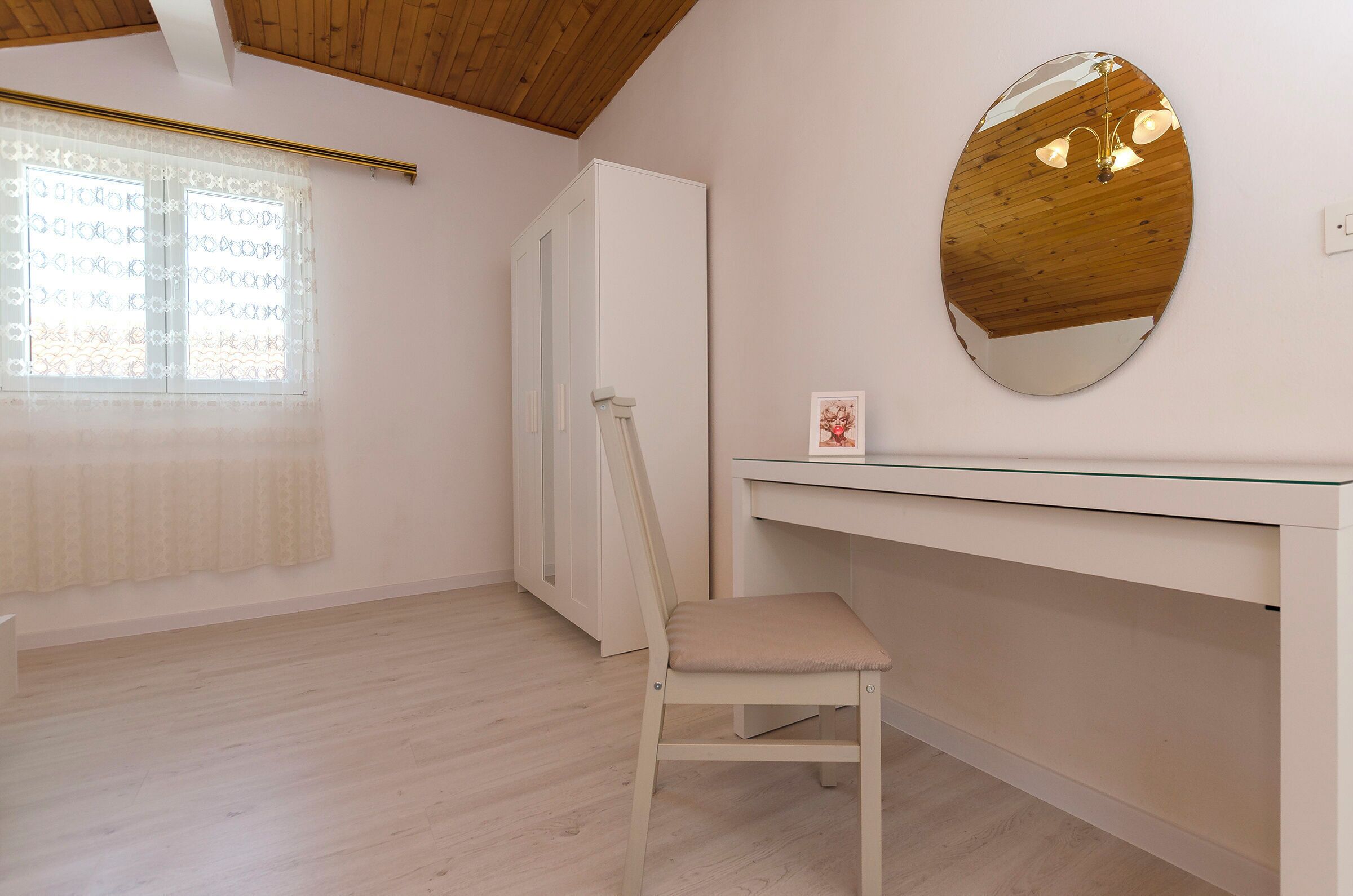Une pièce avec un plafond en bois, un bureau blanc avec un miroir, une chaise, une fenêtre avec des rideaux à motifs et une porte.
