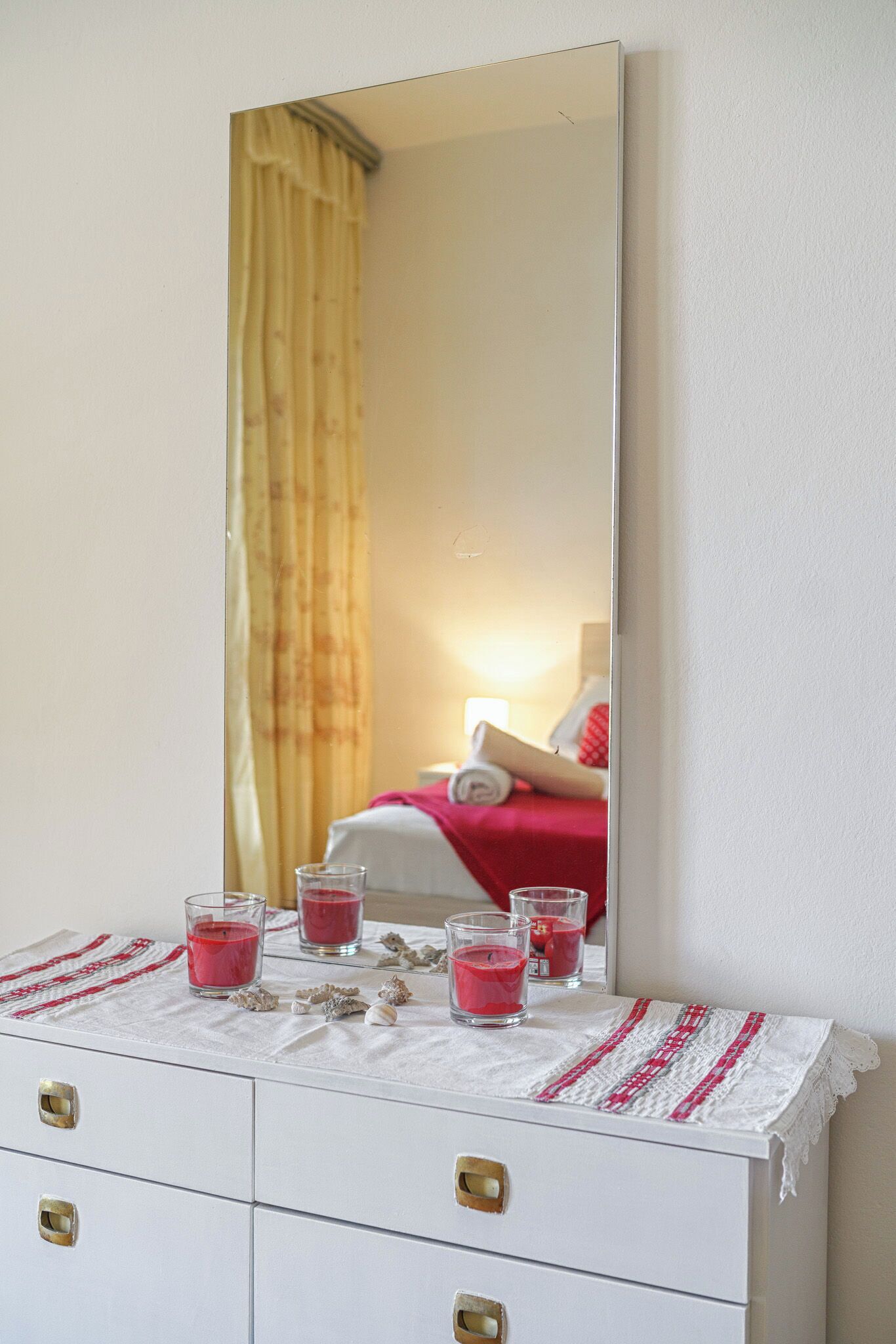 A white dresser with three glass candle holders containing red candles, a white lace tablecloth, and a mirror reflecting a room with a bed and yellow curtains.